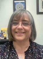 The image shows a woman with shoulder-length hair, smiling for a selfie. She is wearing glasses and a dark top with a colorful pattern. Behind her, there are various decorative items on a shelf, including a small statue, a circular design artwork, and a sign that says "YOU ARE ENOUGH." There is also a peacock feather visible in the background, along with some green plants. The setting appears to be a bright, well-lit indoor space.