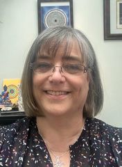 The image shows a woman with shoulder-length hair, smiling for a selfie. She is wearing glasses and a dark top with a colorful pattern. Behind her, there are various decorative items on a shelf, including a small statue, a circular design artwork, and a sign that says "YOU ARE ENOUGH." There is also a peacock feather visible in the background, along with some green plants. The setting appears to be a bright, well-lit indoor space.