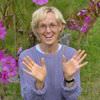 The image depicts a person with light-colored hair, wearing a purple sweater. They are sitting outdoors, smiling and raising their hands in a friendly gesture. The background features vibrant pink flowers and green grass, creating a natural and cheerful setting.