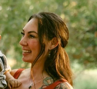 The image features a woman with long, dark hair and visible tattoos, smiling while holding a child. The background appears to be a green, natural setting, suggesting an outdoor environment. The woman is wearing a colorful outfit with a strap, and the child is dressed in a light-colored outfit. The overall atmosphere seems warm and joyful.