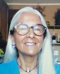 The image shows a woman with long, straight grey hair, wearing large round glasses. She has a warm smile and is dressed in a blue top. In the background, there are shelves with decorative items and plants. The setting appears to be indoors.