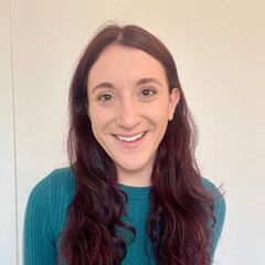 The image shows a person with long, wavy dark hair, wearing a teal ribbed sweater. They are smiling and positioned in a well-lit space with a plain, light background. There is a part of a wall visible on the left side of the image, suggesting an indoor setting.