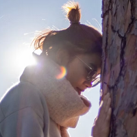 The image depicts a woman with a hairstyle featuring a small bun on top of her head, standing beside a tree. She is wearing sunglasses and has a scarf wrapped around her neck. The background shows a bright light, likely sunlight, creating a halo effect around her head. The woman's head is touching the tree, and the scene captures a serene and contemplative moment.