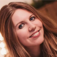 The image features a young person with long, wavy hair and a bright smile. They have blue eyes and appear to be in a well-lit environment, possibly indoors. In the background, there are soft, glowing lights that give a warm ambiance to the scene.