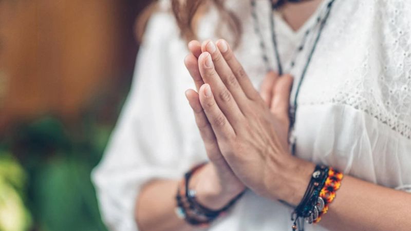 The image shows a close-up of a person's hands placed together in a gesture of prayer or reverence. The person is wearing a white, flowing garment, and there are several bracelets on their wrists. The background suggests a natural or serene environment with greenery visible, giving a calm atmosphere to the scene.