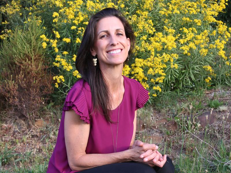 The image shows a person squatting in a grassy area, with a vibrant yellow flowering bush in the background. The person is wearing a purple top and black pants, with black flats on their feet. They are smiling and appear to be enjoying the outdoors. The setting includes various plants and greenery.