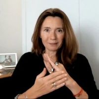 The image shows a woman with long brown hair, sitting in a setting that appears to be an office or a room with light-colored walls. She is wearing a black top and is gesturing with her hands, displaying a specific hand movement. There is a framed photo on a desk behind her, but the contents of the photo are not clear. The overall vibe of the image seems to be professional or personal.