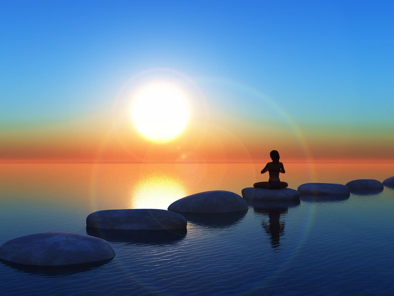 The image depicts a serene scene featuring a calm body of water with smooth, round stones forming a path across it. A silhouette of a person is seated in a meditative pose on one of the stones. The background features a vibrant sunrise, with the sun positioned prominently in the sky, casting warm colors of orange and yellow across the horizon. The overall atmosphere is peaceful and tranquil, evoking a sense of calm and mindfulness.