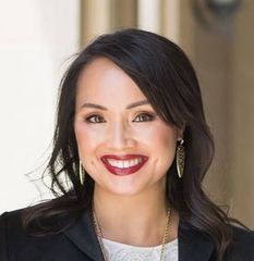 The image shows a woman smiling while standing near an architectural column. She has dark hair and is wearing a black blazer over a white shirt. She accessorizes with a long necklace featuring a blue pendant and tassel. The background appears to be outdoors, with greenery visible, indicating a bright and pleasant environment.