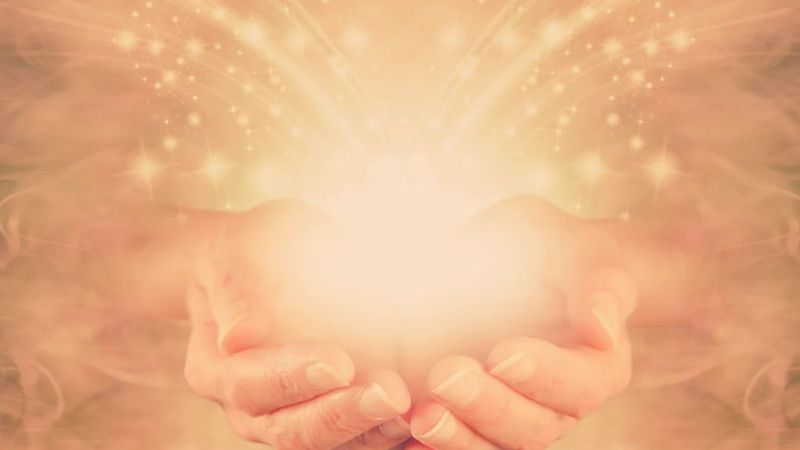 The image features two hands held together in a cupped position, as if cradling something. There is a glowing, bright light emanating from the center of the hands, surrounded by a soft, ethereal glow and sparkles or stars. The background appears hazy and warm in tone, contributing to a mystical or serene atmosphere.