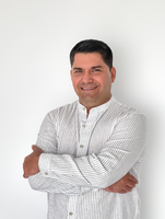 The image features a man with dark hair styled neatly, standing against a plain white background. He is wearing a light-colored, striped shirt and is smiling while crossing his arms. The overall expression is friendly and welcoming.