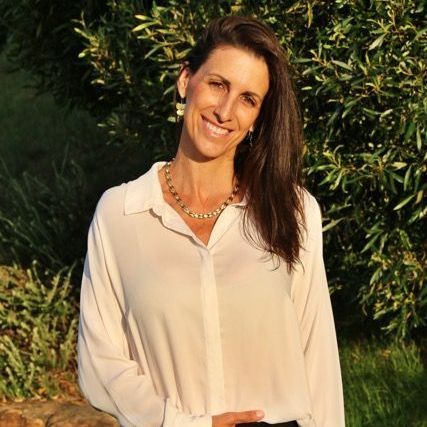 The image features a woman standing outdoors, smiling and posing for the camera. She has long, dark hair and is wearing a light-colored, button-down shirt along with black pants. The background consists of greenery, likely indicating a natural setting, with some rocks visible on the ground. The lighting appears warm, suggesting it may be late afternoon or early evening.